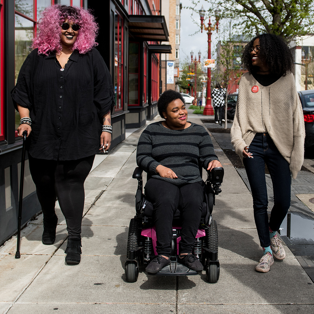 blind woman with cane and woman in wheelchair going down sidewalk with another woman all smiling